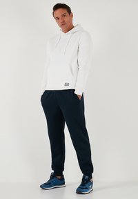 Witte katoenen hoodie met een voorvak en trekkoord. Marineblauwe joggingbroek met elastische boorden en zijzakken. Blauwe sneakers met zwarte accenten.