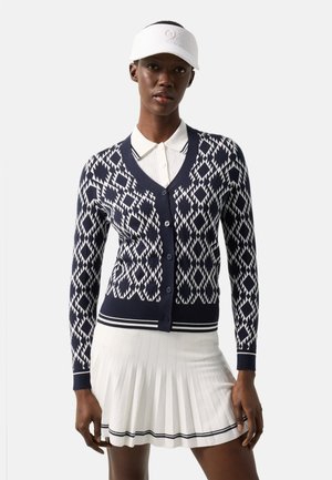 Frau trägt eine marineblaue und weiße Strickjacke mit geometrischem Muster, ein weißes Hemd mit Kragen, einen plissierten weißen Rock und ein weißes Schildmützchen vor einem einfarbigen Hintergrund.