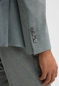 Close-up van de hand van een persoon naast de manchet van een grijze colbert met vier donkere knopen op de mouw.