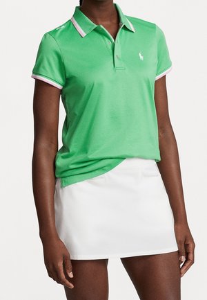 Frau trägt ein grünes kurzärmeliges Poloshirt mit pinkem Besatz und weißem Logo, kombiniert mit einem weißen Rock.