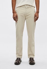 Μπεζ βαμβακερό παντελόνι με ίσια γραμμή, εμπρόσθιες τσέπες και κλείσιμο με κουμπί, σε συνδυασμό με καφέ δερμάτινα παπούτσια, με λεία υφή.