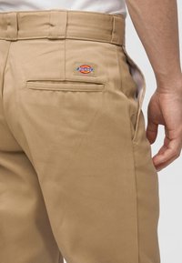 Close-up van de beige Dickies broekzak op een persoon die een wit overhemd draagt, met zichtbare ceintuurlussen en een hand vlakbij de zijnaden.