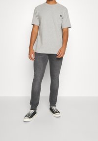 Camiseta gris de manga corta, con un bolsillo en el pecho. Lleva puesta con jeans oscuros ajustados y zapatillas negras con suelas blancas.