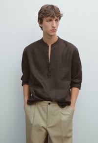 Brown linen shirt with a collarless design, long sleeves rolled up, and three button closures. Paired with beige pleated trousers.