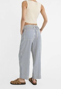Persoon in blauw en witte verticaal gestreepte wijde broek, crèmekleurige mouwloze crop top en bruine sandalen, staand met handen in de zakken.