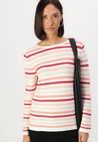 Striped long-sleeve top featuring red, pink, peach, and beige lines on a cream background, made of a ribbed fabric. Black shoulder bag included.