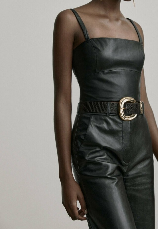 Femme portant un top sans manches en cuir noir et un pantalon taille haute assorti, avec une ceinture noire texturée et une grande boucle dorée.