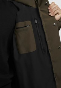 Padded Jacke mit einem dunklen schwarzen Innenfutter und einem olivgrünen Äußeren, ausgestattet mit einer Reißverschlusstasche auf der Brust und Druckknopfdetails.