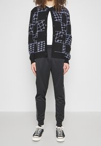 Svart cardigan med lila geometriskt mönster, vit skjorta under, kombinerad med svarta byxor med sidostripes och svarta sneakers.