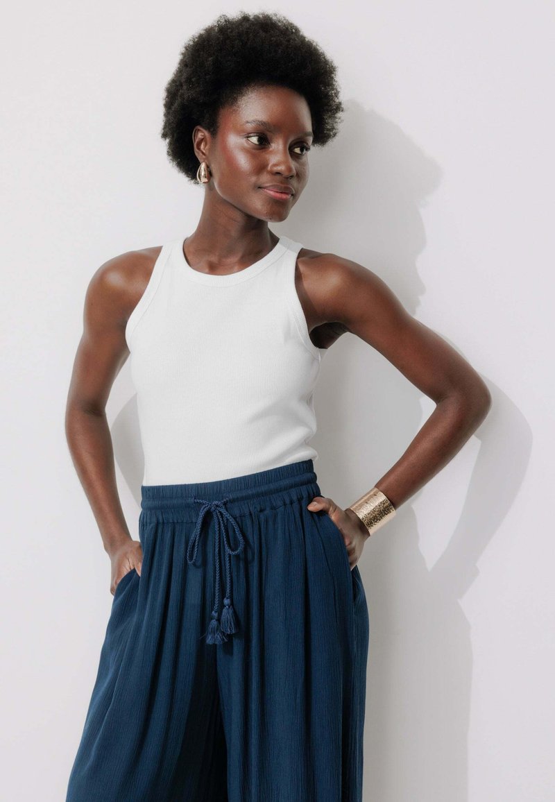 Haut sans manches blanc en maille côtelée associé à un pantalon large plissé bleu marine, avec une taille à cordon de serrage et un bracelet à clairaut en or.