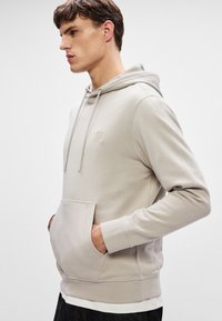 Vaalea beige huppari, jossa on kiristettävä huppu, kengurupussi, ribbaus hihansuissa ja pieni logo vasemmassa yläkulmassa. Pehmeä kangas.