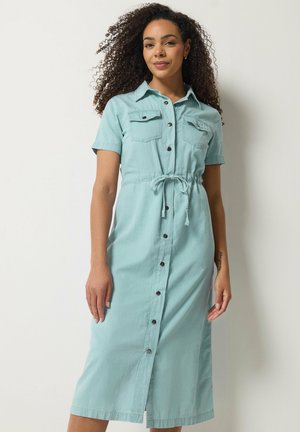 Kobieta o kręconych włosach, ubrana w jasnoniebieską sukienkę midi zapinaną na guziki z krótkimi rękawami i wiązaniem w talii, stojąca na jednolitym białym tle.