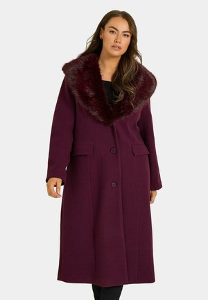 Femme portant un long manteau violet foncé avec un grand col en fausse fourrure, une fermeture à boutons et des poches à rabats, debout devant un fond uni.