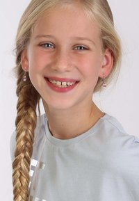 Zilveren oorbellen met een roze cupcake ontwerp op een meisje met lang gevlochten blond haar, dat een lichtblauwe t-shirt draagt en glimlacht.