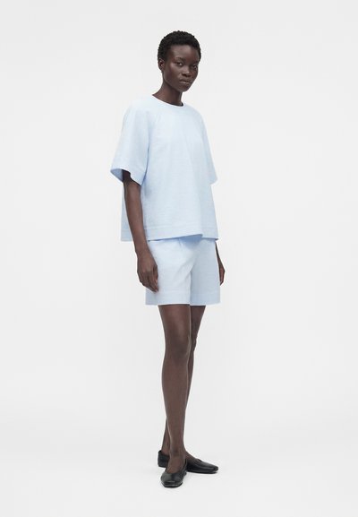 Γυναίκα που στέκεται φορώντας ανοιχτό μπλε κοντομάνικο τοπ και αντίστοιχα σορτς με μαύρα επίπεδα παπούτσια σε απλό λευκό φόντο.