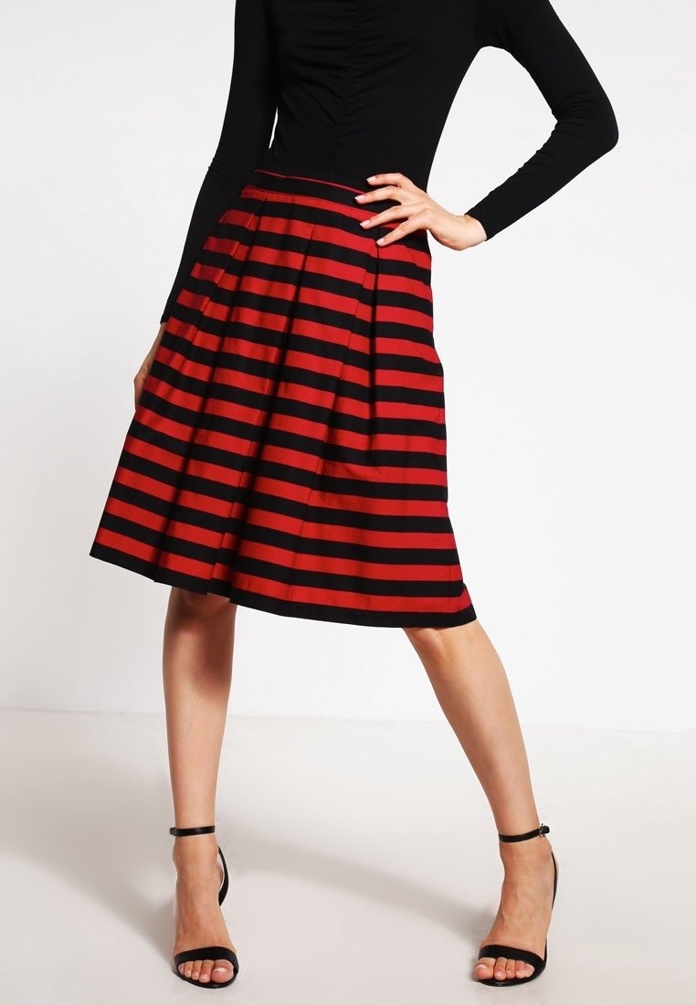 Femme portant une jupe mi-longue rayée rouge et noire à rayures horizontales, un haut noir à manches longues et des sandales à talons hauts noires à brides autour de la cheville.