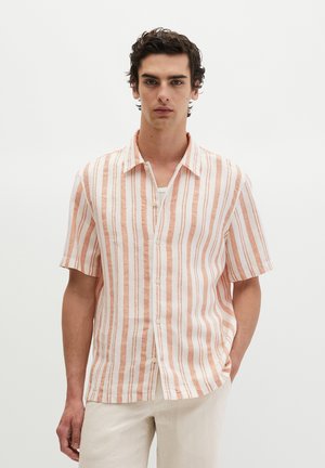 Junger Mann mit dunklem lockigem Haar, der ein aufgeknöpftes kurzärmeliges beiges und weißes vertikal gestreiftes Hemd und helle Hosen trägt, Hände in den Taschen.