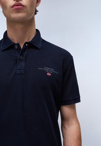 Mężczyzna ubrany w granatową koszulkę polo z guzikami i logo Napapijri z małą norweską flagą na piersi.