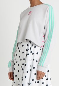 Felpa corta grigio chiaro con strisce verde menta e logo Adidas rosa, abbinata a una gonna bianca con pois neri.