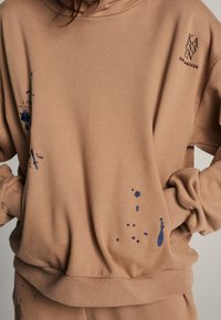 Sweatshirt marron avec capuche, texture douce et éclaboussures de peinture bleue, avec un petit logo sur la poitrine.