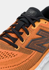 Orange atletik sko med en tekstureret mesh-overdel, sorte snørebånd og et stort sort logo. Har letvægtsmaterialer og gummi sål accenter.