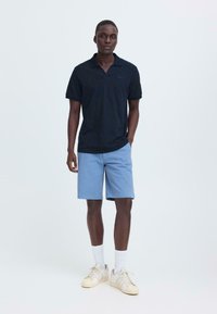 Polo bleu marine à manches courtes associé à un short bleu clair. Le modèle porte des chaussettes blanches et des baskets crème, debout devant un fond uni.