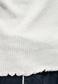 Εγγύς πλάνο από ριμπ ύφασμα πλεκτό σε εκρού χρώμα με κυματιστό τελείωμα πάνω από σκούρο παντελόνι.