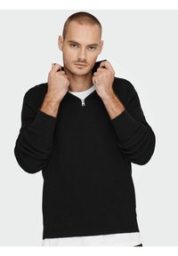 Suéter negro acanalado con cuello con cremallera, de mangas largas y un ajuste relajado. Llevado sobre una camisa blanca, mostrando una tela texturizada.