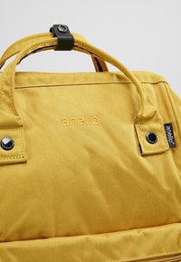 Sac à dos en tissu jaune avec deux poignées au sommet, arborant un logo en relief et des accents en métal noir. Texture lisse et design minimaliste.