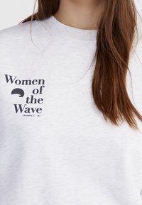 Sudadera blanca con el texto "Mujeres de la Ola" y un logo de ola en el lado izquierdo del pecho, llevada por una persona con cabello largo y castaño.