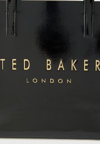 Fekete lakk kézitáska kidomborodó arany betűkkel: "TED BAKER" és "LONDON". Sima textúra, masszív fogantyúk és strukturált forma.