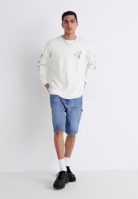 Valkoinen pitkähihais shirt grafiikkasuunnittelulla, yhdistettynä sinisiin farkkushortsaihin. Mustat kengät ja valkoiset sukat täydentävät rentoa ilmettä.