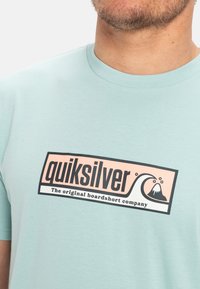 Svetle modré bavlněné tričko s výrazným černým a růžovým logem "Quiksilver", se grafikou vln a hor, krátkými rukávy a kulatým výstřihem.
