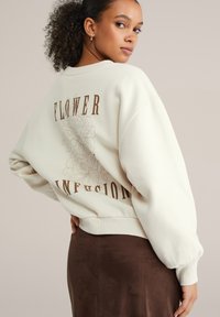 Frau mit lockigem Haar, die einen beigen Pullover mit dem Aufdruck "FLOWER INFUSION" und floralem Design auf dem Rücken trägt, kombiniert mit einem braunen Rock.