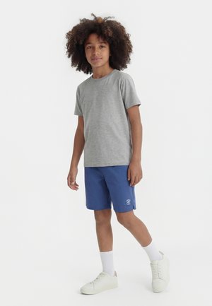 Copil cu păr creț purtând un tricou gri, pantaloni scurți albaștri, șosete albe și adidași, stând pe un fundal alb simplu.