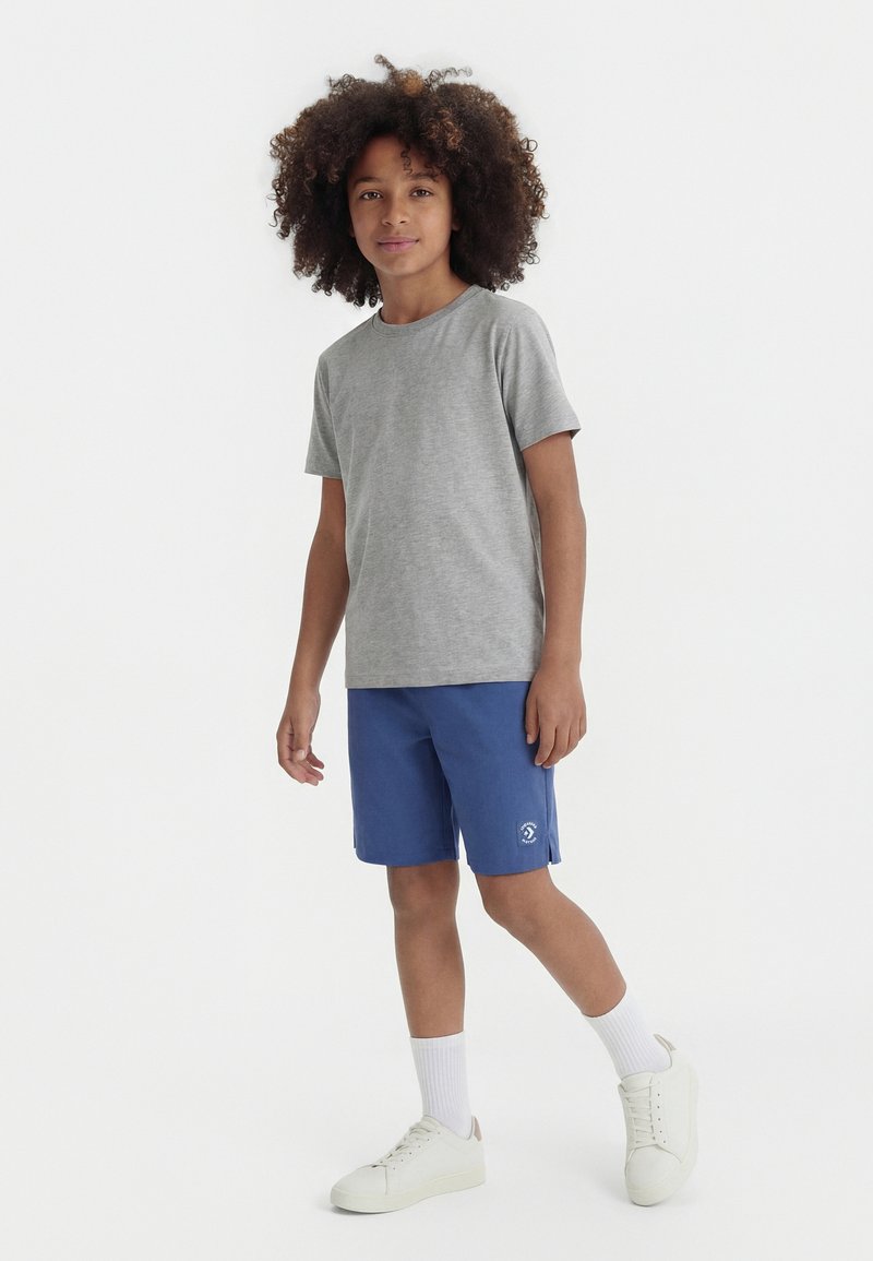 Παιδί με σγουρά μαλλιά που φορά γκρι μπλουζάκι, μπλε σορτς, λευκές κάλτσες και αθλητικά παπούτσια, στέκεται μπροστά σε απλό λευκό φόντο.