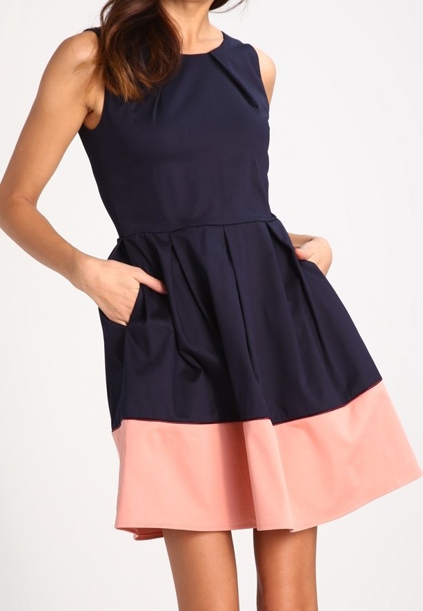 Marineblauwe mouwloze jurk met een geplooid rokje, voorzien van zijzakken en een roze zoom. Gemaakt van een gladde stof met een ronde halslijn.