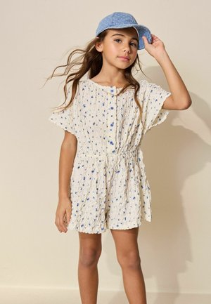 Mädchen trägt einen weißen, strukturierten Overall mit blauem Blumendruck und Knöpfen, richtet eine blaue Jeansmütze, steht vor einem schlichten beigen Hintergrund.