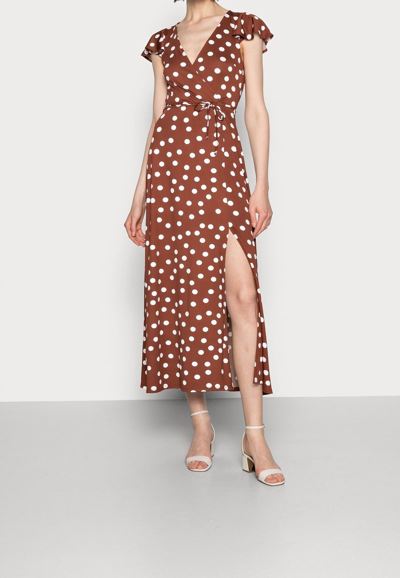 Femme portant une robe portefeuille marron à pois blancs, manches volantées, fente latérale et sandales blanches à talons blocs avec bride cheville.