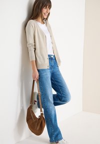 Femme en cardigan beige, chemise blanche, jean bleu, appuyée contre un mur, tenant un sac à bandoulière en daim marron, portant des baskets blanches.