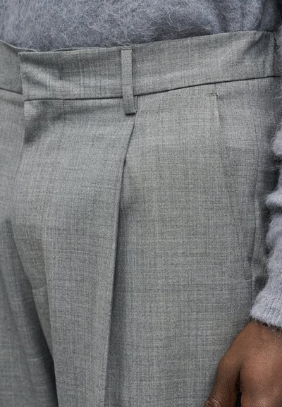 Κοντινό πλάνο ενός ατόμου που φοράει γκρι παντελόνι ραμμένο στα μέτρα και γκρι πουλόβερ με υφή, δείχνοντας την υφή του υφάσματος και ένα χαλαρό χέρι στο πλάι τους.