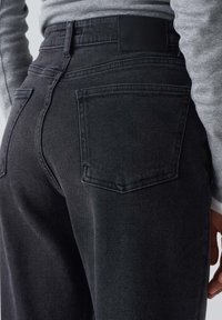 Vaqueros de mezclilla negros con un diseño de pierna recta, que cuentan con dos bolsillos traseros y una parche de cuero en la cinturilla. Detalle de tela texturizada visible.