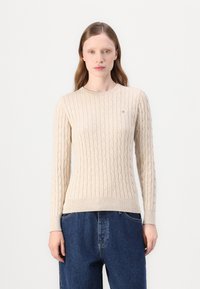 Pulover bej din tricot cu model de cablu, cu guler rotund, mansete și tiv cu modalità, având un mic logo pe piept. Textură moale, design ajustat.