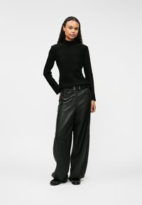 Mustapohjainen, korkeakauluksinen ribbipaita, jossa on diagonaalinen kuvio, yhdistettynä leveälahkeisiin mustiin nahkahousuihin ja matalapohjaisiin mustiin kenkiin. Yksinkertainen, moderni muotoilu.