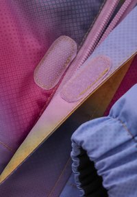 Färgglad tyg i rosa och blå graderingar med prickiga mönster, med dragkedja och kardborrestängningar, samt samlade elastiska ärmslut.