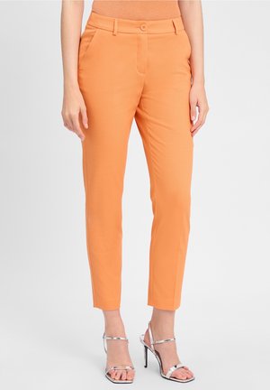 Nainen, joka pukeutunut kapealahkeisiin nilkkapitkiin oransseihin housuihin, joissa on sivutaskut ja napillinen vyötärönauha, parina hopeiset korkeakorkoiset sandaalit.