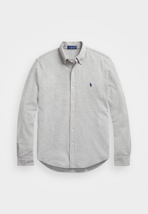 Camisa de botão cinza de manga longa com colarinho, apresentando um pequeno logotipo azul-marinho no lado esquerdo do peito e uma bainha curvada. Feita de tecido suave.