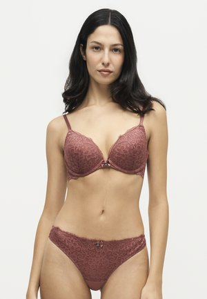 Mulher com cabelo comprido e escuro a usar um conjunto coordenado de sutiã e roupa interior em renda malva, em pé contra um fundo claro simples.