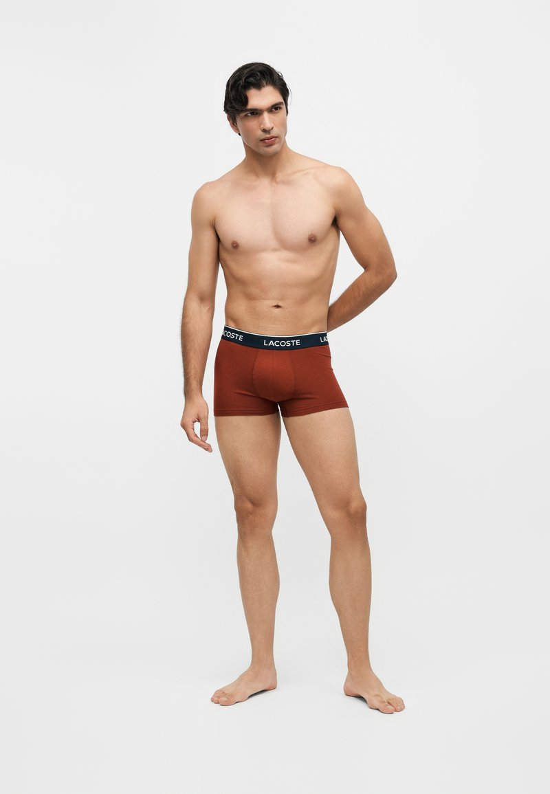 Homme torse nu aux cheveux foncés portant un boxer couleur rouille avec une ceinture noire Lacoste, debout devant un fond blanc uni.