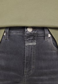 Gros plan sur la ceinture d'un jean gris foncé avec un bouton en métal et une petite étiquette de marque, associé à un haut en tissu côtelé vert olive.